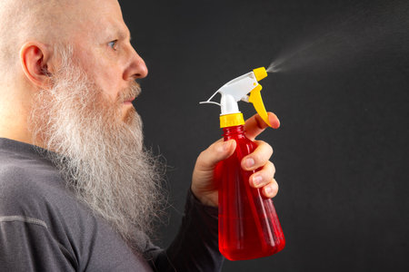 bald and bearded man splashes liquid from a spray bottle on a dark gray background
