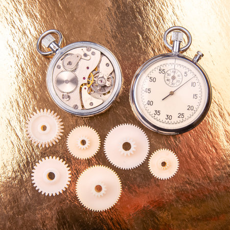 A vintage stopwatch rests alongside intricate clock gears on a reflective golden surface. The...