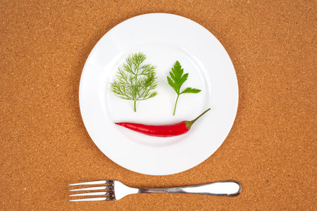 A white plate is creatively arranged with a red chili pepper, cilantro, and dill, forming a smiling...