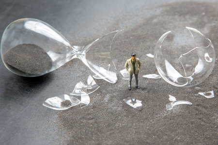 A miniature person poses near a broken hourglass and scattered shards of glass. The dark surface...