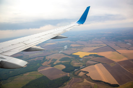 Airplane wing extends from the side as it flies above patchwork fields of green, brown, and gold....