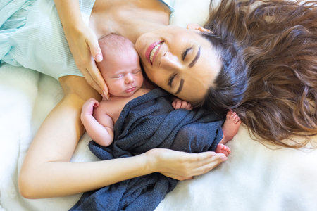 A mother holds her sleeping baby close, smiling gently as they relax on a soft surface. The warm...