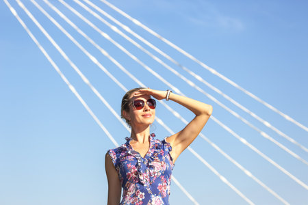 A woman stands on a bridge, dressed in a floral outfit and sunglasses, shielding her eyes from the sun.の写真素材