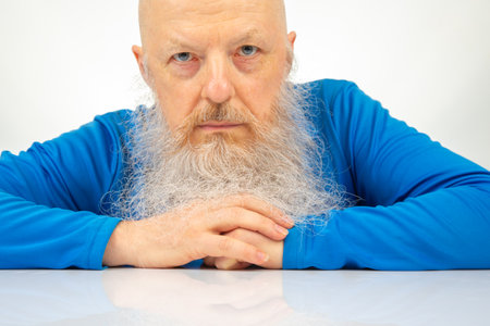A man with a long white beard sits at a table, wearing a bright blue shirt. He gazes seriously at the camera with a focused expression. Natural light illuminates the minimal indoor space.の写真素材