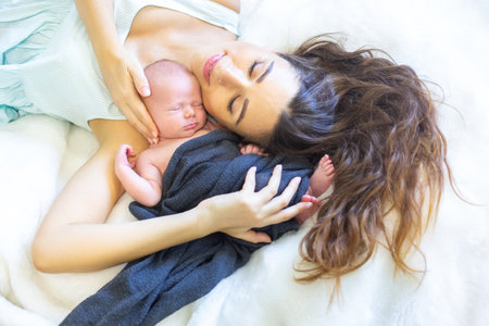 A mother lies on soft bedding, cradling her newborn baby close to her chest. The intimate moment reflects warmth and love as both peacefully rest together in their cozy environment.の写真素材