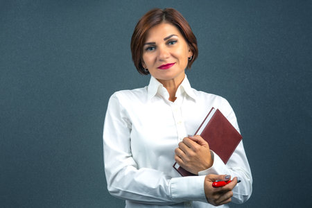 A woman wearing a white shirt stands confidently, holding a book in one arm and a pen in the other. She displays a friendly smile, with a dark gray backdrop adding to the professional ambiance.の写真素材