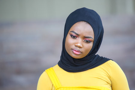 An African woman wearing a black hijab and a yellow top appears to be deep in thought at an outdoor...