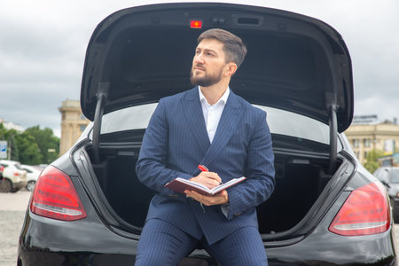A businessman dressed in a dark suit sits on the trunk of a black car, taking notes in a notebook. He looks thoughtful while a cityscape with cloudy skies is visible in the background.の写真素材