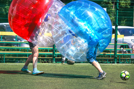 Two players in large inflatable bubbles collide while playing bubble soccer on a green turf field. The sun shines brightly, creating an energetic atmosphere for the fun sport.の写真素材