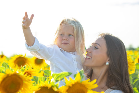 A mother holds her child high while they explore a field full of sunflowers on a bright, sunny day. The child playfully gestures, and both share a joyful moment in nature's beauty.の写真素材