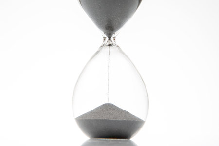 An hourglass filled with black sand is positioned upright, showing the steady flow of sand from the top chamber to the bottom, highlighting the passage of time in minimalist style.の写真素材