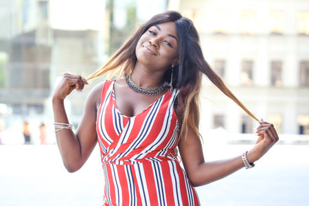 Confident African woman stands outside, smiling and enjoying the moment while styling her long hair....