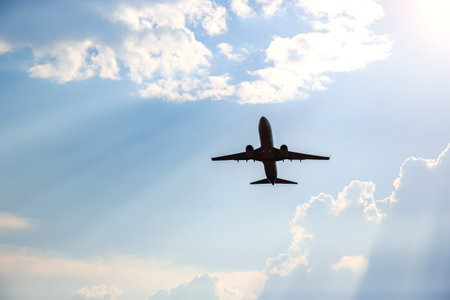 An airplane ascends into the azure sky filled with soft, fluffy clouds while the sun casts warm rays across the horizon during late afternoon.の写真素材