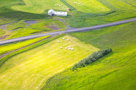 Lush green fields stretch endlessly, framing a white farmhouse on an isolated plot in Iceland, with scattered bales in the foreground under a bright sky during daytime.の写真素材