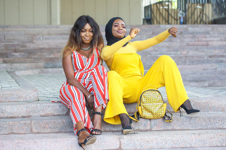 Two African women are sitting on stone steps, confidently posing and smiling. One is wearing a striped outfit and the other a bright yellow jumpsuit.の写真素材