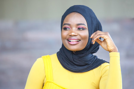 An African woman wears a yellow outfit and a black hijab while smiling and enjoying the outdoors. Her joyful expression reflects confidence and poise against a neutral background.の写真素材