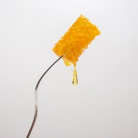 A piece of golden honeycomb is suspended from a stainless steel fork, with honey slowly dripping down. The background is light and neutral, emphasizing the rich color and texture of the honeycomb.の写真素材