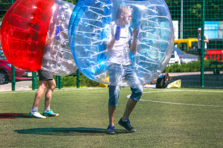 Two players participate in bubble soccer, wearing inflatable spheres while enjoying a vibrant day on an artificial turf field. The atmosphere is lively with bright colors and energetic movement.の写真素材