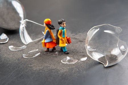 Two miniature people walk cautiously past broken glass pieces scattered on a dark surface, creating a dramatic contrast. Their vibrant clothing adds color to the somber environment.の写真素材