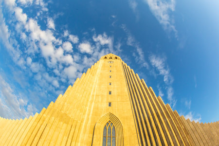 Hallgrimskirkja church stands tall with its distinctive design, bathed in sunlight against a backdrop of vibrant blue sky and scattered clouds, showing Reykjavik's architectural beauty.の写真素材