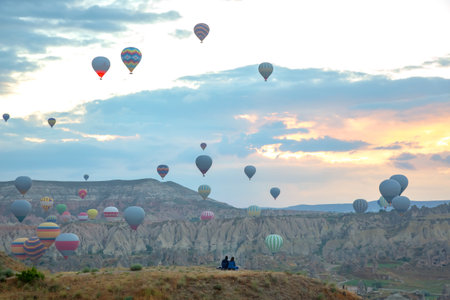 Visitors admire the colorful hot air balloons soaring above the unique rock formations of Cappadocia, Turkey, as the sun rises, casting a beautiful light over the landscape.の写真素材