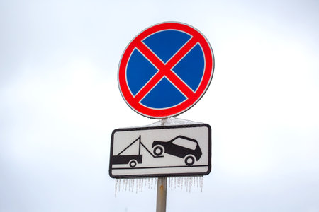 A no parking sign with a blue circle and red slash is displayed, accompanied by a towing warning. Icicles hang from the sign in a cold, overcast winter setting, indicating freezing conditions.の写真素材