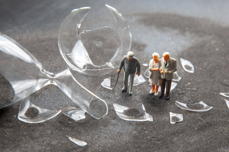 Miniature elderly people stand near broken glass remnants on a textured surface, observing the scattered shards as they interact. The setting conveys a quiet moment of reflection amidst chaos.の写真素材