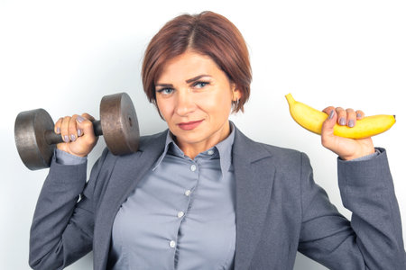 A businesswoman in a gray suit holds a dumbbell in one hand and a banana in the other, showing a blend of fitness and healthy eating. The light backdrop emphasizes her confident expression.の写真素材