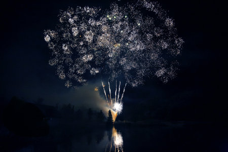 Bright fireworks explode in vibrant colors above a serene lake, reflecting light on the water's surface during a joyful summer festival. Trees silhouette against the illuminated sky.の写真素材