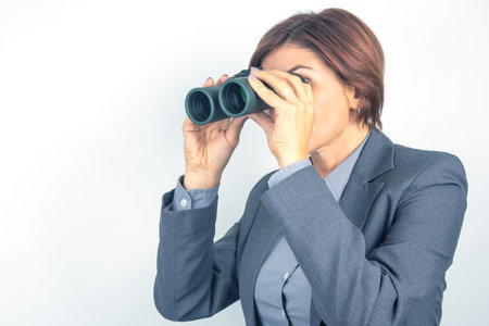 A businesswoman in a gray suit focuses intently while using binoculars, possibly scouting for opportunities or analyzing a distant view in her workspace.の写真素材