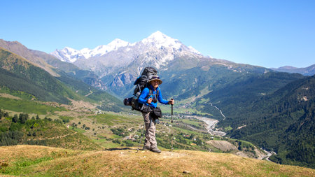 A hiker enjoys the breathtaking view from a mountain summit, surrounded by majestic snow-capped peaks. The clear blue sky enhances the beauty of the lush green valley below during daytime.の写真素材