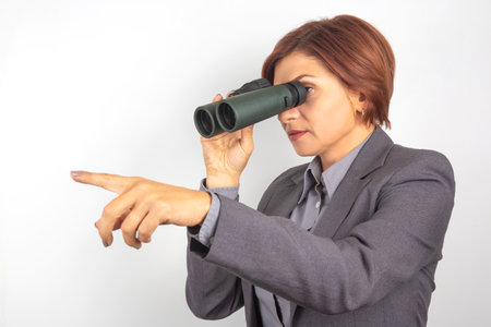A professional woman in a suit peers through a pair of binoculars, focused on a distant target. She points ahead, demonstrating curiosity and determination in her exploration effort.の写真素材