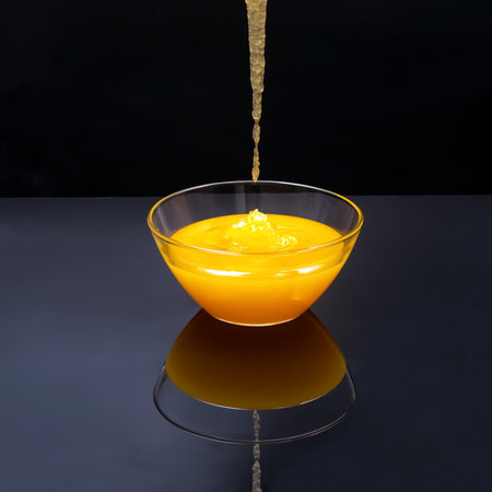 Thick golden syrup pours smoothly into a clear glass bowl, creating a beautiful reflection on a shiny black surface. The syrup's rich color contrasts vividly with the dark background.の写真素材