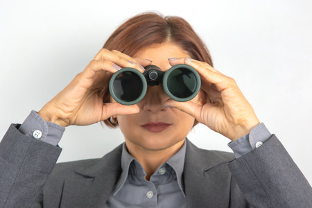 A businesswoman in a gray suit examines her environment through binoculars, showcasing a focused expression. The setting suggests a professional atmosphere with neutral background colors.の写真素材