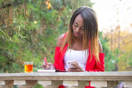 An African woman stands at a stone table in a park, wearing a vibrant red jacket. She is writing notes in a notebook and sipping tea from a glass, surrounded by beautiful autumn foliage.の写真素材