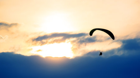 A paraglider gracefully glides above fluffy clouds as the sun sets, illuminating the sky with warm hues. The serene atmosphere captures the beauty of adventure in evening light.の写真素材
