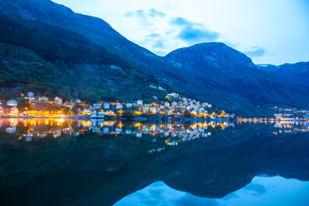 A charming village in Norway sparkles with lights as dusk settles. The serene water reflects colorful houses and the mountains loom in the background, creating a tranquil atmosphere.の写真素材