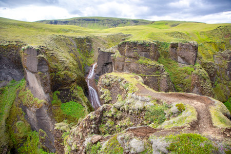 A breathtaking view of a canyon featuring a cascading waterfall. Rich green vegetation blankets the cliffs, showing the stunning natural beauty of Iceland on a cloudy day.の写真素材