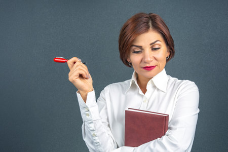 A confident woman stands against a neutral background, holding a notebook in one arm while pondering something with a writing pen in her other hand, showing a moment of reflection at work.の写真素材