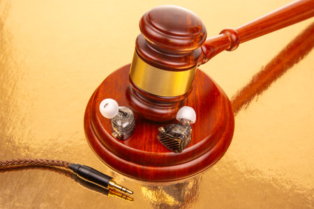 A wooden gavel rests on a shiny golden surface alongside high-quality earphones and a cable, creating a striking contrast. The focus is on the blend of legal symbolism and audio technology.の写真素材