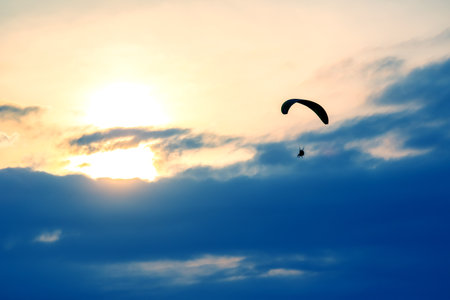 A skydiver gracefully descends through the clouds at sunset, surrounded by a stunning array of colors. The silhouette of the parachute contrasts against the vibrant sky as daylight fades.の写真素材