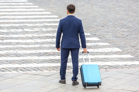 Businessman in a suit waits at a crosswalk holding a blue suitcase, preparing to navigate city streets on a bright day, blending professionalism and travel.の写真素材