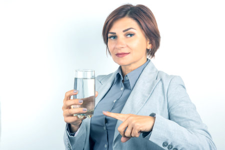 A professional woman with short brown hair in a gray suit holds a clear glass of water. She points confidently with her right finger, suggesting importance. The setting appears clean and modern.の写真素材