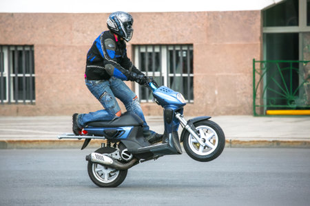 A rider in protective gear skillfully performs a wheelie on a blue scooter on a bustling city street. The sun shines brightly, highlighting the rider's balanced posture and determination.の写真素材