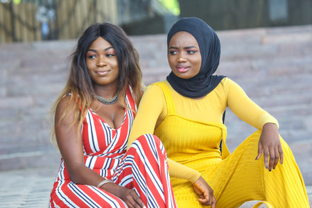 Two African women are seated together in a lively outdoor location, wearing bright and stylish...