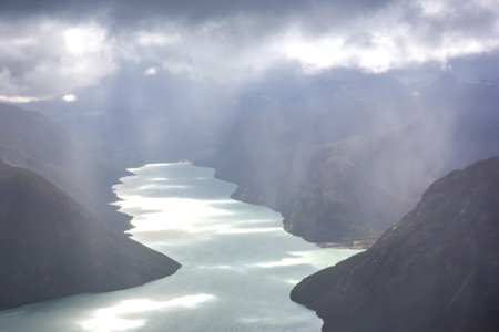 Majestic fjords of Norway showcase stunning views where calm waters reflect the sky. Clouds create a mystical atmosphere as light breaks through, illuminating the serene landscape.の写真素材