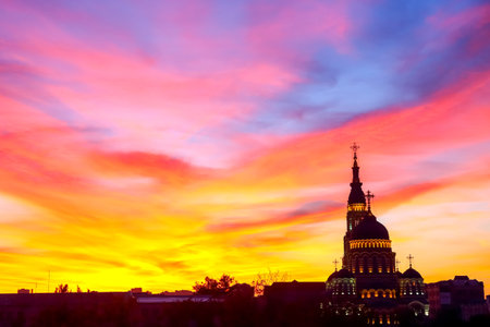 Vibrant colors fill the sky at sunset as a church silhouette stands tall against the backdrop of the urban horizon, creating a stunning contrast with the deep hues of purple, orange, and pink.の写真素材