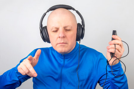 A man with a bald head wearing headphones closes his eyes in concentration, holding a music device in one hand while he enjoys the rhythm of his favorite tunes in a calm indoor setting.の写真素材