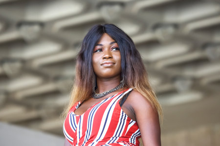 A woman of African descent stands proudly, showing her fashion sense in a vibrant atmosphere. The background features a modern architectural design, highlighting a sunny day ambiance.の写真素材