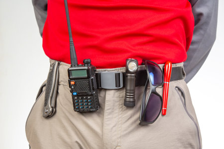 A person stands with a red shirt and gray pants, showcasing a radio, flashlight, and sunglasses attached to their belt. This setup suggests readiness for outdoor activities or emergencies.の写真素材